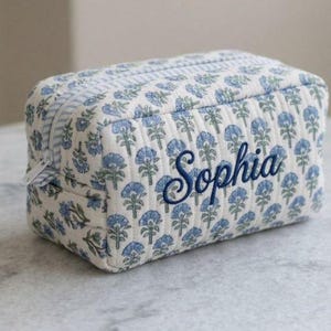 Op de afbeelding: Een rechthoekige, doorgestikte toilettas met een lichtblauw en groen bloemenpatroon op een witte achtergrond. De naam Sophia is in donkerblauw cursief schrift op de voorzijde geborduurd. Een lichtblauw en wit gestreepte ritssluiting is zichtbaar.