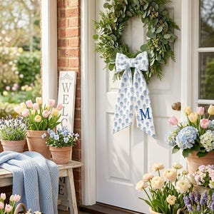 K&ouml;nnte beinhalten: Ein gr&uuml;ner Kranz mit einer blau-wei&szlig;en, floral gemusterten Schleife mit dem Buchstaben M, der an einer wei&szlig;en T&uuml;r h&auml;ngt. Ein Holzschild tr&auml;gt die Aufschrift WELCOME, ebenso wie eine braune Fu&szlig;matte. T&ouml;pfe mit Tulpen, Hortensien und Lavendel.