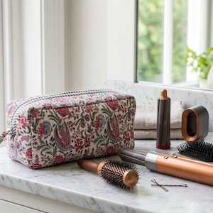 May include: A floral patterned cosmetic bag with a black and white striped zipper, hair styling tools, and a hairbrush on a marble countertop. The bag features red, blue, and green floral designs.