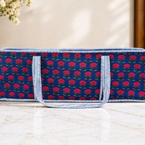 May include: A rectangular quilted fabric tote bag featuring a deep blue background with a repeating pattern of small pink flowers and green stems. The bag is finished with white and blue striped fabric trim along the edges and two matching handles.
