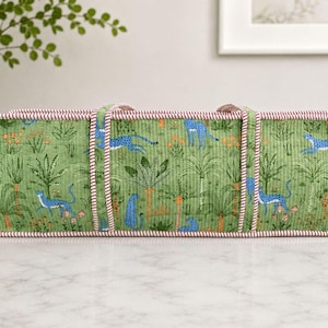 May include: A rectangular quilted fabric storage bin featuring a green background with a repeating pattern of blue leopards, palm trees, and orange flowers. The bin has two fabric handles and is finished with a red and white striped trim.