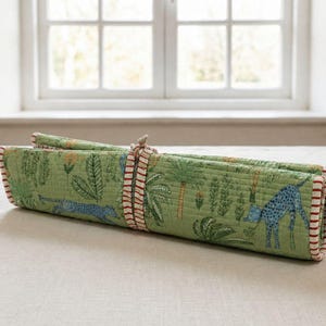 May include: A rolled quilted fabric mat featuring a green background with a repeating pattern of blue spotted animals, palm trees, and orange flowers. The mat is secured with a matching fabric tie featuring thin red and white horizontal stripes.