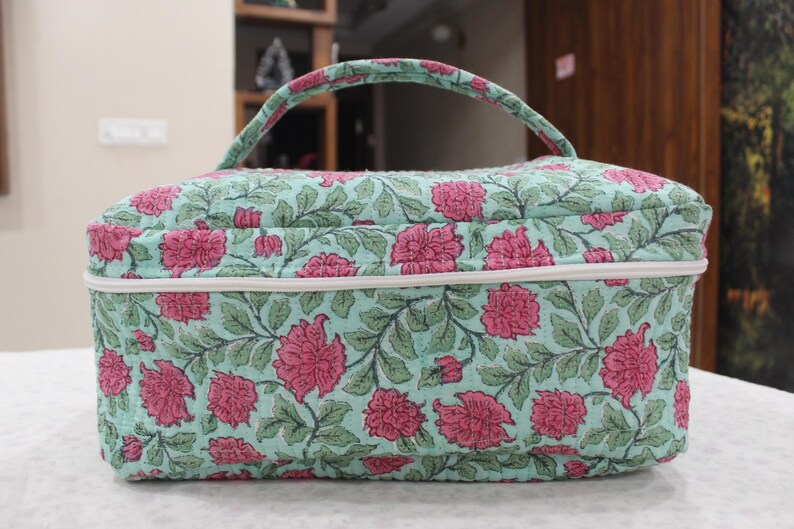 May include: A rectangular cosmetic bag with a handle, featuring a floral pattern of pink flowers and green leaves on a light blue background. The bag has a white zipper and trim.