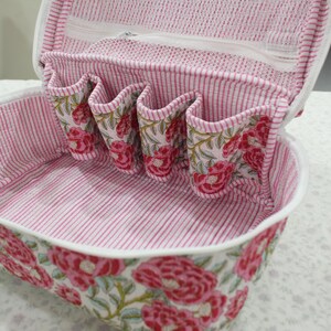 May include: Open cosmetic bag with a floral pattern. The bag is white with a pink and red floral design on the exterior. The interior features pink and white striped fabric and several small pockets.