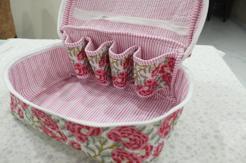 May include: Open cosmetic bag with a floral pattern. The bag is white with a pink and red floral design on the exterior. The interior features pink and white striped fabric and several small pockets.