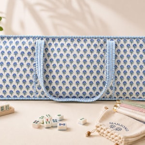 May include: A rectangular quilted carrying case with a blue and green floral pattern on a white background, featuring blue and white striped trim and handles. Beside it are scattered mahjong tiles and a beige drawstring bag labeled Mahjong.