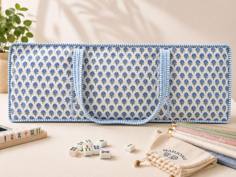May include: A rectangular quilted carrying case with a blue and green floral pattern on a white background, featuring blue and white striped trim and handles. Beside it are scattered mahjong tiles and a beige drawstring bag labeled Mahjong.