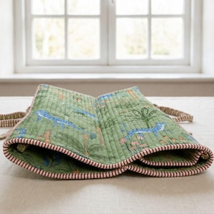 May include: A quilted green fabric roll featuring a repeating pattern of blue cheetahs and palm trees. The edges are finished with a red and white striped trim, and the piece includes matching striped fabric ties on the sides for securing the roll.