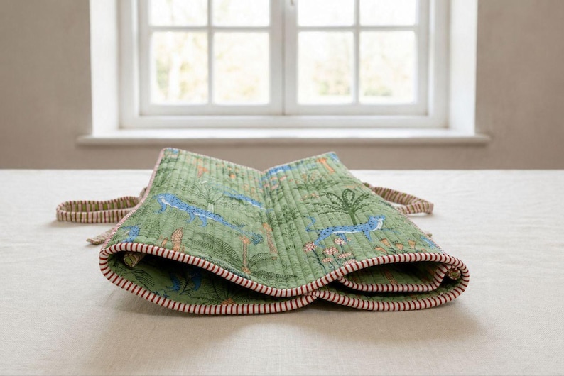 May include: A quilted green fabric roll featuring a repeating pattern of blue cheetahs and palm trees. The edges are finished with a red and white striped trim, and the piece includes matching striped fabric ties on the sides for securing the roll.