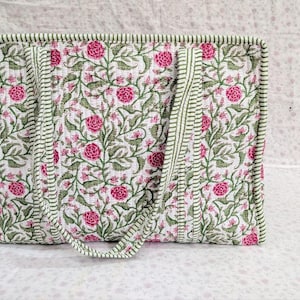 May include: A rectangular quilted tote bag featuring a white background with a repeating pattern of pink flowers and green leafy vines. The bag includes two matching fabric handles and is finished with green and white striped fabric trim.