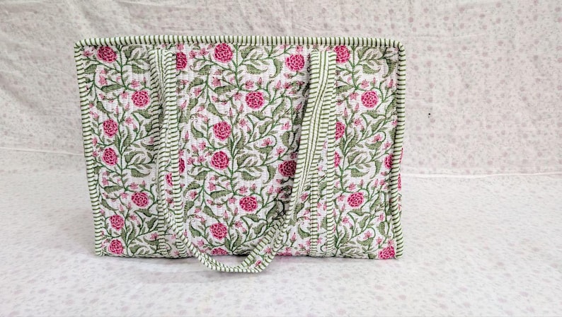 May include: A rectangular quilted tote bag featuring a white background with a repeating pattern of pink flowers and green leafy vines. The bag includes two matching fabric handles and is finished with green and white striped fabric trim.