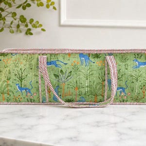 May include: A rectangular quilted fabric storage bin featuring a green background with a repeating pattern of blue leopards, palm trees, and orange flowers. The bin has two fabric handles and is finished with red and white striped piping trim.