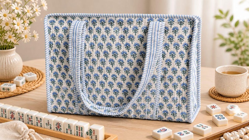 May include: A rectangular quilted tote bag featuring a repeating blue and green floral block print pattern on a white background, finished with light blue and white striped fabric trim along the edges and two matching striped shoulder straps.