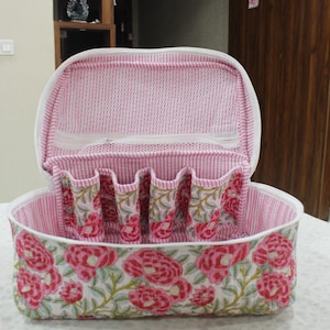 May include: Open, floral-patterned cosmetic bag with a white zipper and handle. The bag features a pink and white striped interior and a white exterior with a large pink floral design. The bag has several small pockets inside.