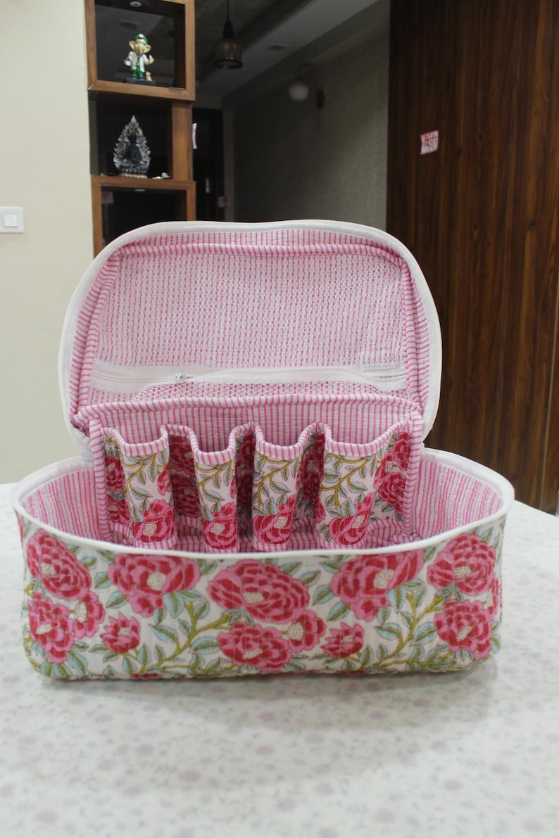 May include: Open, floral-patterned cosmetic bag with a white zipper and handle. The bag features a pink and white striped interior and a white exterior with a large pink floral design. The bag has several small pockets inside.