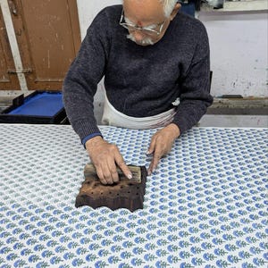 May include: A rectangular wooden block with a carved floral pattern used for textile printing, pressed onto a white fabric surface featuring a repeating blue and green floral motif. The block has a natural dark wood finish and a handle on top.