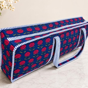 May include: A rectangular quilted fabric bag featuring a deep blue background with a repeating pattern of pink flowers and green stems. The bag has a zippered top closure and is finished with white and blue striped fabric trim and matching handles.