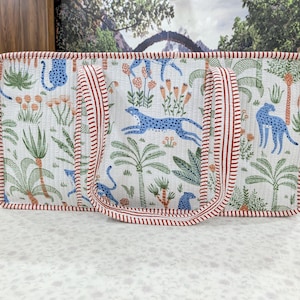 May include: A rectangular quilted tote bag featuring a white background with a repeating pattern of blue cheetahs, green palm trees, and orange flowers. The bag has two red and white striped fabric handles and matching red and white striped trim.