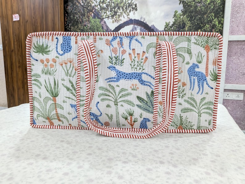 May include: A rectangular quilted tote bag featuring a white background with a repeating pattern of blue cheetahs, green palm trees, and orange flowers. The bag has two red and white striped fabric handles and matching red and white striped trim.