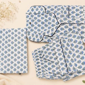 Op de afbeelding: Een witte pyjamaset met een herhalend blauw en groen bloemenpatroon in blokdruk. De set bestaat uit een overhemd met lange mouwen en knopen met zwarte biezen, een bijpassende broek en een gevouwen stoffen tasje met hetzelfde motief.