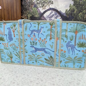 May include: A rectangular quilted tote bag in light blue featuring a repeating pattern of blue leopards, green palm trees, and orange floral accents. The bag has two vertical fabric handles and is finished with a green and white striped trim.