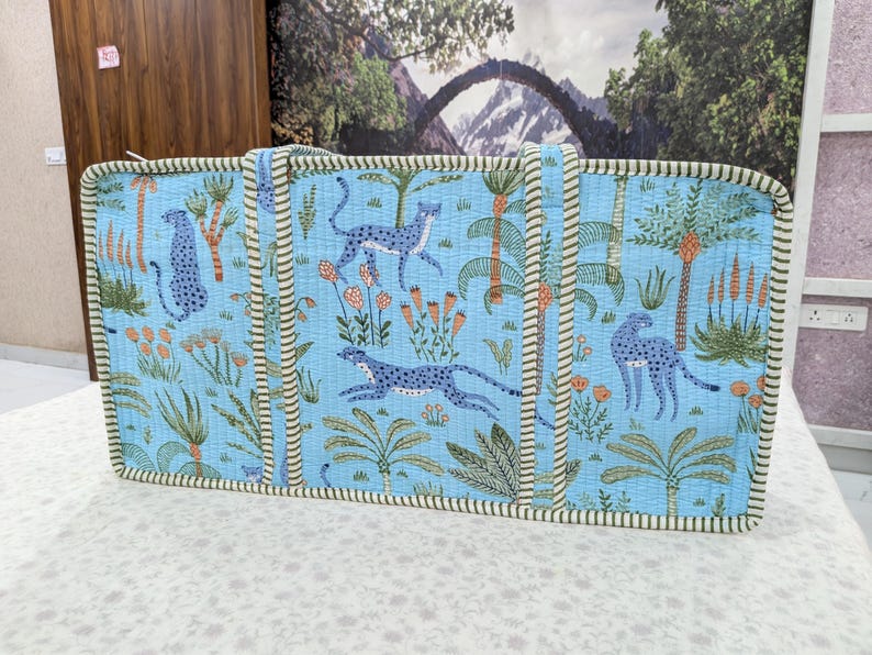 May include: A rectangular quilted tote bag in light blue featuring a repeating pattern of blue leopards, green palm trees, and orange floral accents. The bag has two vertical fabric handles and is finished with a green and white striped trim.