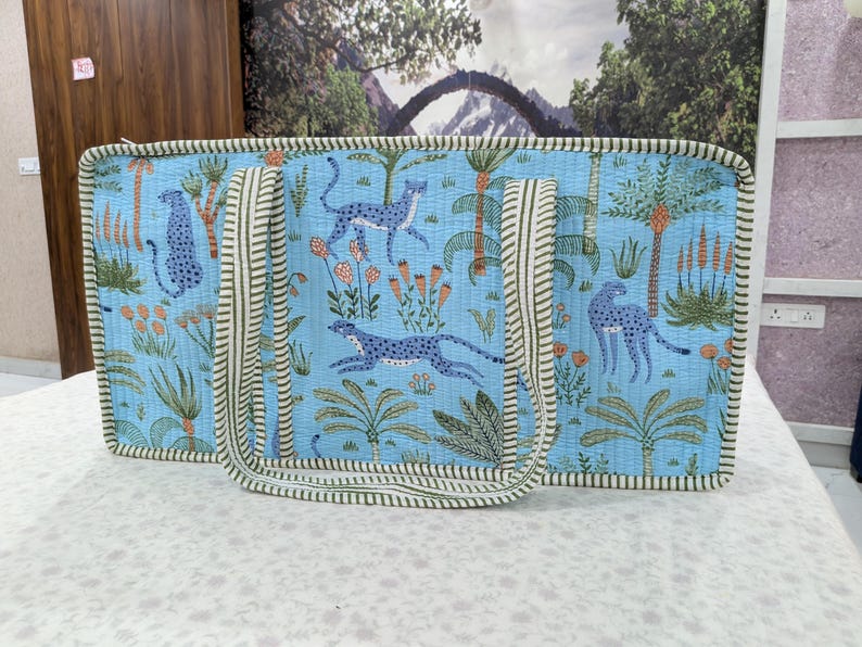 May include: A rectangular quilted tote bag in light blue featuring a repeating pattern of dark blue cheetahs, palm trees, and orange flowers. The bag has two long, striped fabric handles and matching striped piping along all the outer edges.