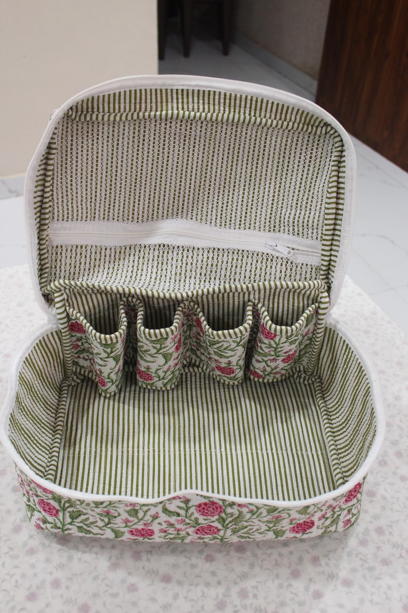 May include: Open, white-trimmed cosmetic bag with a floral print on the exterior and a green and white striped interior. The bag has a zippered top and several small pockets inside for organizing items.