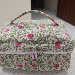 May include: A floral-patterned cosmetic bag with a white background, pink flowers, and green leaves. The bag has a white zipper, a handle, and a quilted texture. The bag is approximately 10 inches wide and 8 inches tall.