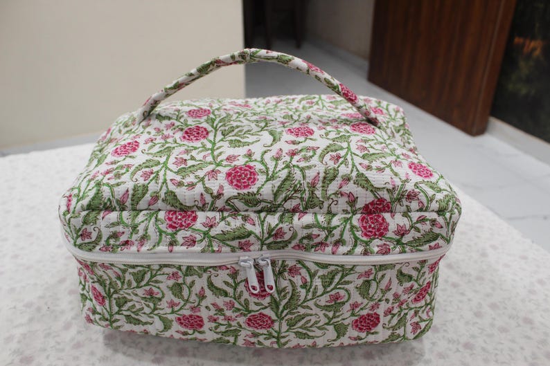 May include: A floral-patterned cosmetic bag with a white background, pink flowers, and green leaves. The bag has a white zipper, a handle, and a quilted texture. The bag is approximately 10 inches wide and 8 inches tall.