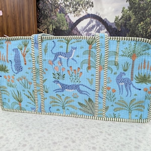May include: A rectangular quilted tote bag in light blue featuring a repeating pattern of blue leopards, green palm trees, and orange floral accents. The bag has two vertical fabric handles and is finished with a green and white striped trim.