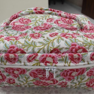 May include: A floral-patterned cosmetic bag with a handle. The bag features a white background with a repeating pattern of pink flowers and green leaves. It has a white zipper closure and a matching floral handle.