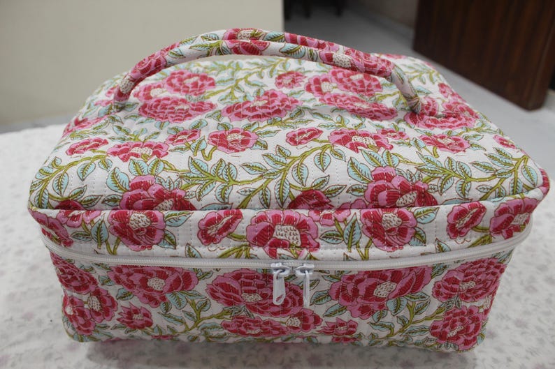 May include: A floral-patterned cosmetic bag with a handle. The bag features a white background with a repeating pattern of pink flowers and green leaves. It has a white zipper closure and a matching floral handle.