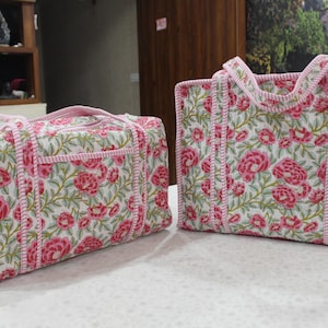 May include: Two quilted fabric bags with a floral design. The bags have a white base with pink roses and green leaves. The edges and handles are trimmed with pink fabric. One bag is rectangular, and the other is a tote bag.