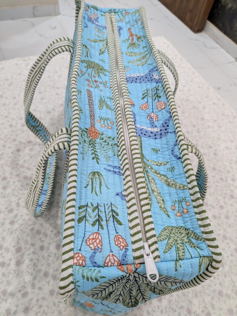 May include: A light blue quilted tote bag featuring a repeating botanical print of green leaves, orange flowers, and blue abstract shapes. The bag has a white zipper closure and is finished with green and white striped fabric trim and two handles.