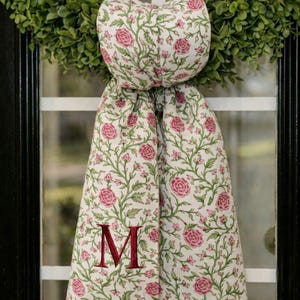 May include: A floral fabric door swag with a green wreath in the background. The swag features a white background with pink roses and green vines. A burgundy letter "M" is embroidered on the fabric.