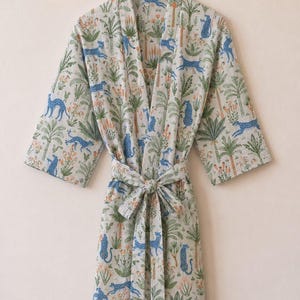 May include: A light-coloured robe with a jungle-themed pattern. The robe features blue leopards, palm trees, and floral accents. It has a matching belt and is hanging on a wooden hanger.