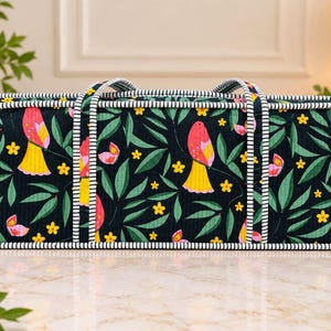 May include: A rectangular storage bag with a black background featuring a repeating pattern of pink and yellow birds, green leaves, and small yellow flowers. The bag has two handles and is trimmed with a white and navy blue striped fabric border.