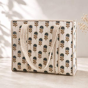 Cream quilted mahjong tote bag with brown and navy floral block print, striped padded handles, and structured spacious design, displayed in a neutral minimalist setting with soft natural light and decorative vase.