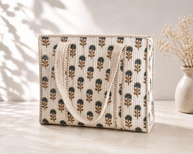 Cream quilted mahjong tote bag with brown and navy floral block print, striped padded handles, and structured spacious design, displayed in a neutral minimalist setting with soft natural light and decorative vase.