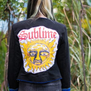 May include: A black jacket featuring a large, circular patch on the back with a yellow sun graphic and the word Sublime in red gothic lettering. The jacket has long sleeves with blue cuffs and a blue hemline, worn over dark gray denim shorts.