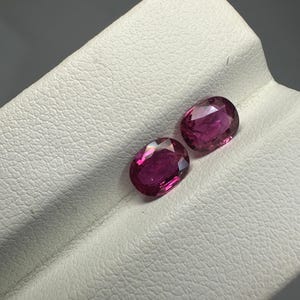 Puede incluir: Dos gemas ovaladas de color rosa intenso se exhiben sobre una superficie blanca texturizada. Las gemas tienen un aspecto facetado, captando la luz y mostrando su color vibrante. El fondo es de un gris suave.