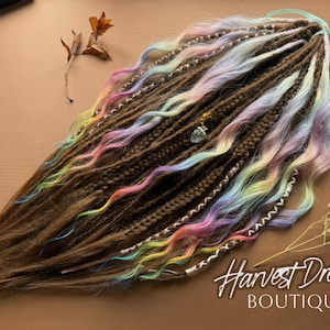 Puede incluir: Dreadlocks marrones con reflejos de arcoíris pastel y acentos ondulados. Las rastas tienen algunos pequeños amuletos y están dispuestas sobre una superficie marrón. El texto "Harvest Dreads BOUTIQUE" es visible.