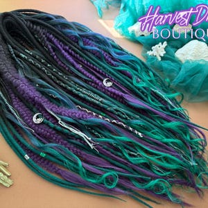 May include: A set of synthetic dreadlocks in shades of black, teal, and purple. The dreads are adorned with silver accents and charms. The image includes the text "Harvest Dreads Boutique".