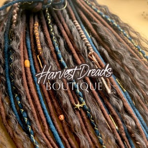 Puede incluir: Primer plano de rastas en tonos marrón, gris y azul. Algunas rastas están decoradas con cuentas y detalles dorados. El texto "Harvest Dreads Boutique" es visible.