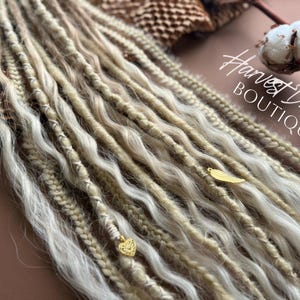 May include: Close-up of blonde and light brown dreadlocks with gold-colored feather and heart charms. The dreadlocks are wavy and textured, with a mix of light and dark tones. The text "Harvest Dreads Boutique" is visible in the upper right corner.
