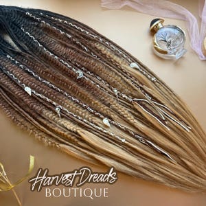 May include: Ombre dreadlocks in shades of black, brown, and blonde, adorned with silver accents. The hair extensions are styled with decorative silver wire and charms. The Harvest Dreads Boutique logo is visible.