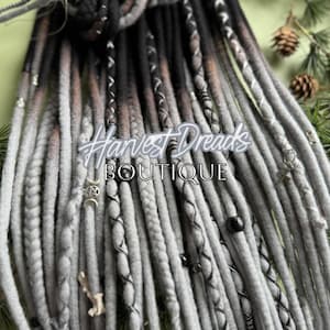 May include: Close-up of a bundle of gray, black, and brown dreadlocks. The dreadlocks are decorated with small charms and beads. The text "Harvest Dreads BOUTIQUE" is visible in the center of the image.