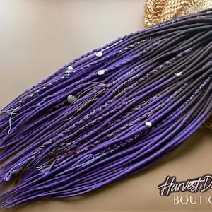 May include: A set of synthetic dreadlocks in various shades of purple, ranging from deep violet to bright lavender. The strands feature braided sections, wrapped accents, and small white shell beads. The text Harvest Dreads BOUTIQUE is visible.