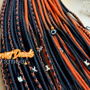 May include: Close-up of dreadlocks in black and orange. The dreads are decorated with orange stitching, small charms, and beads. The text "Harvest Dreads Boutique" is in the image. The dreadlocks are a hair accessory.